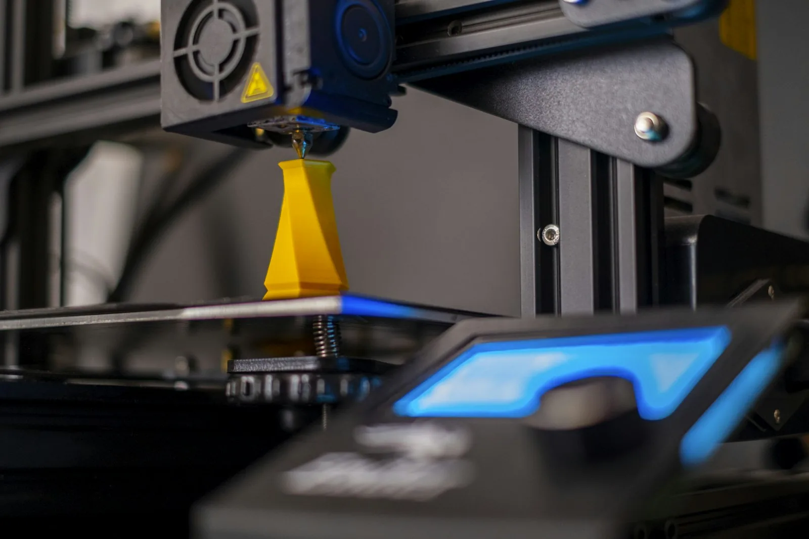 Bico da impressora 3D depositando camadas em peça amarela, com brilho azul